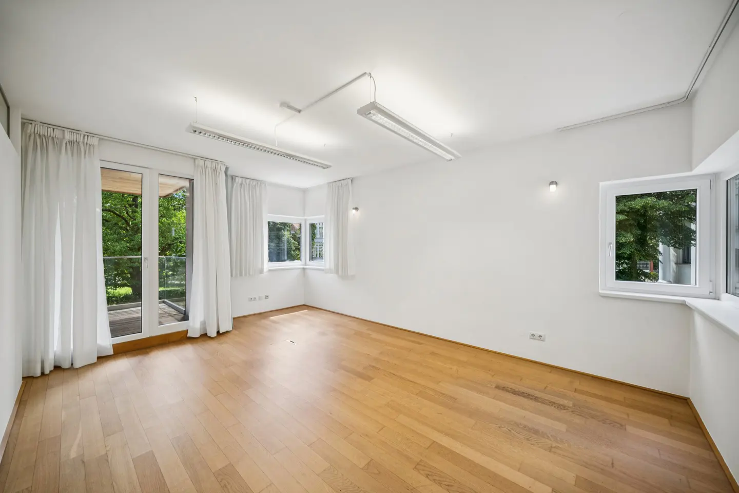Bright, empty room with white walls, light wood floors, large windows with white curtains, and modern ceiling lighting.