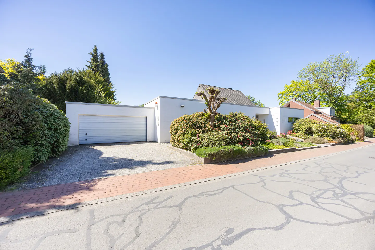 Modern white house with a gray garage door, green bushes, and a gray driveway on a sunny day.