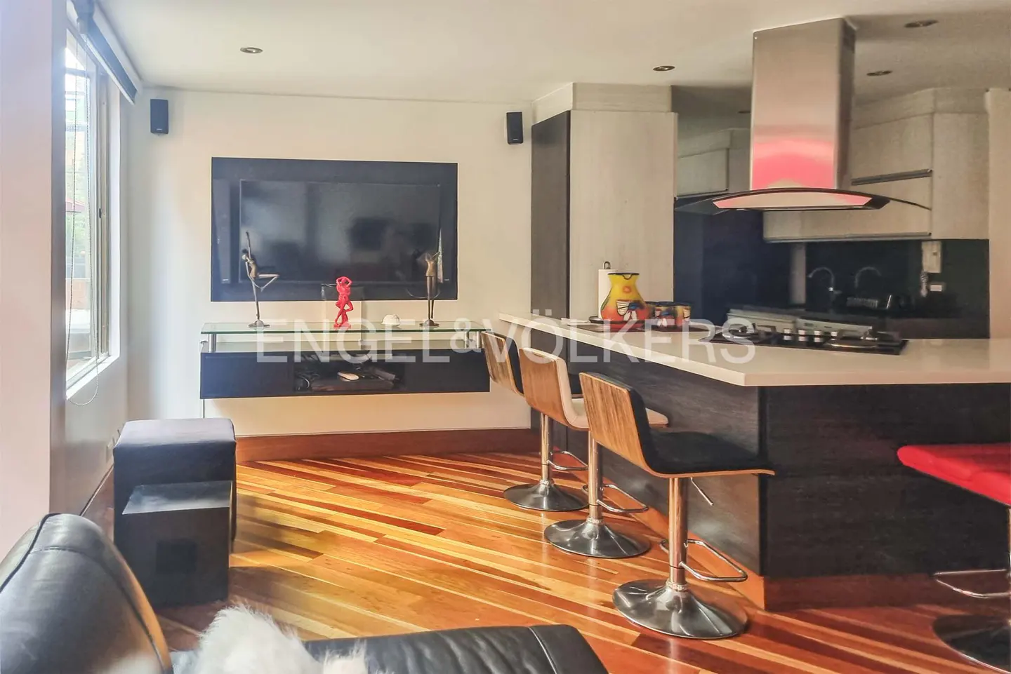 A modern kitchen and living area with wood floors, bar stools at the counter, and a TV on the wall.