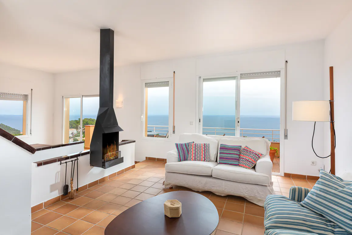 Bright living room with a modern black fireplace, white sofa with patterned pillows, and a stunning ocean view.