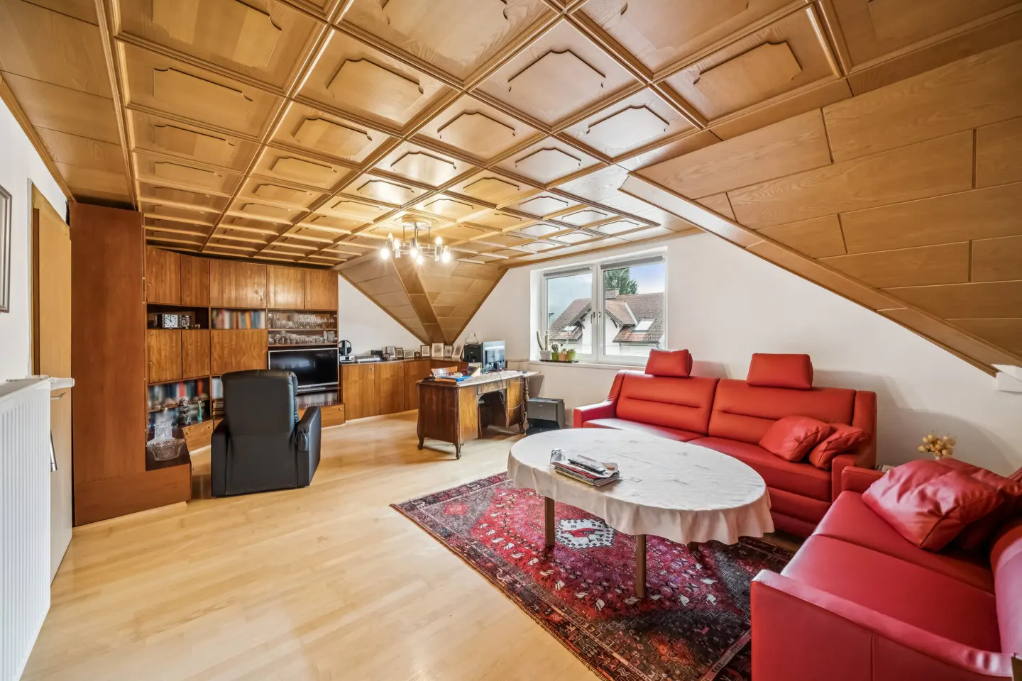 Attic living room with wood-paneled ceiling, red sofas, desk, and built-in shelving. A patterned rug sits beneath a white-clothed table.