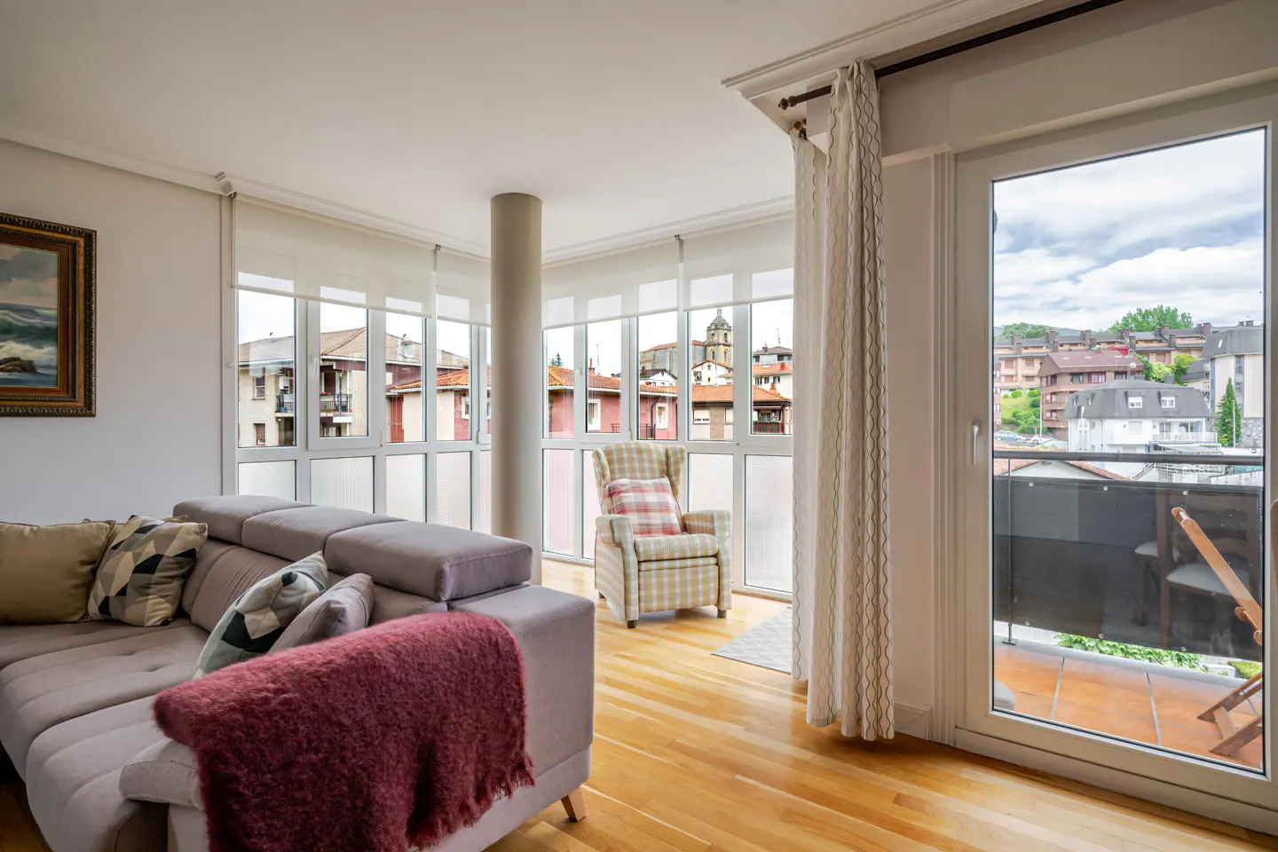 Bright living room with a grey sofa, plaid armchair, hardwood floors, and a balcony overlooking a town.