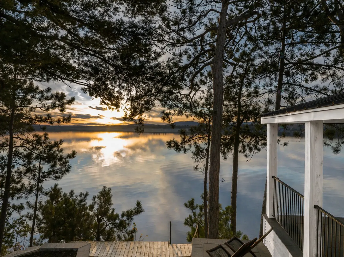 Lake view from a wooden deck with trees. Sunset reflects on the water. White porch with black railing on the right.