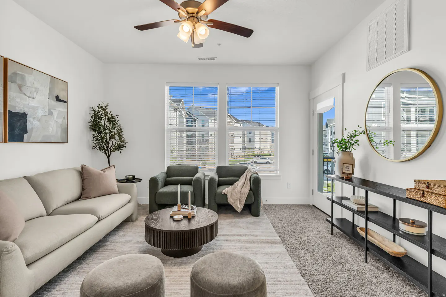 Bright living room with a beige sofa, two green armchairs, and a round coffee table on a gray rug. A ceiling fan hangs above.