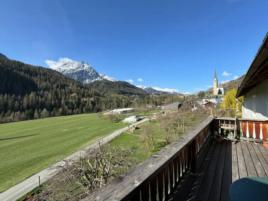 View from a wooden balcony overlooking a green field, village, and snow-capped mountains under a clear blue sky.