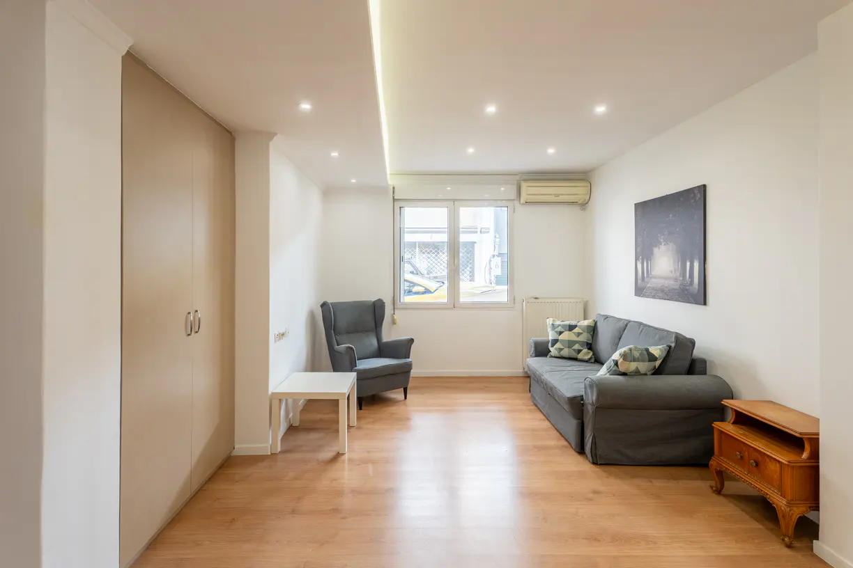 Bright living room with wood floors, white walls, and recessed lighting. Features a gray sofa, armchair, and a small wooden cabinet.