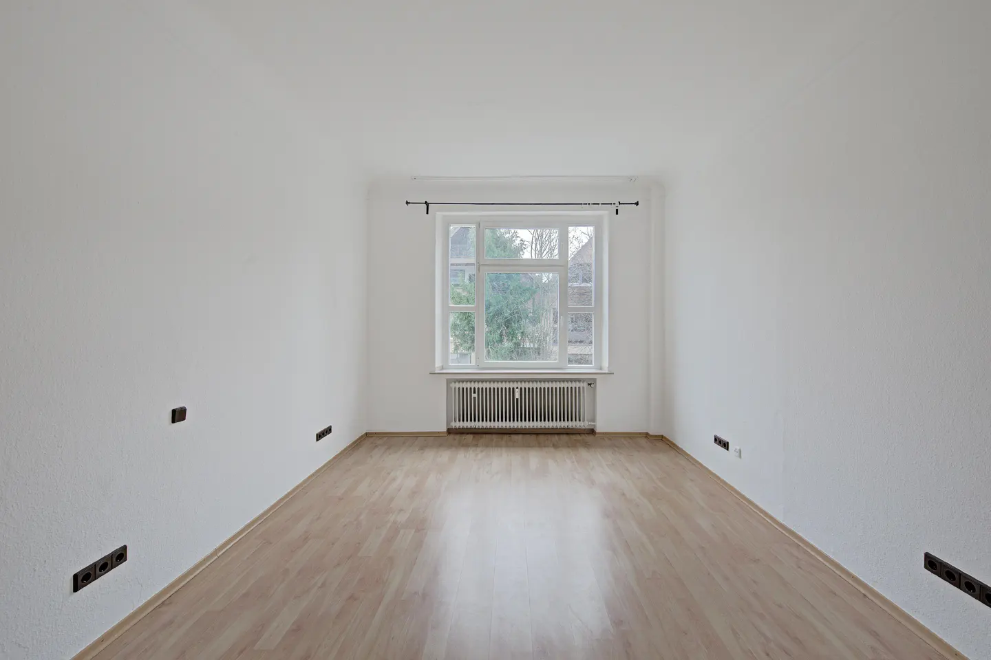 Empty room with light wood floor, white walls, and a window with a radiator below it. Electrical outlets are visible on the walls.