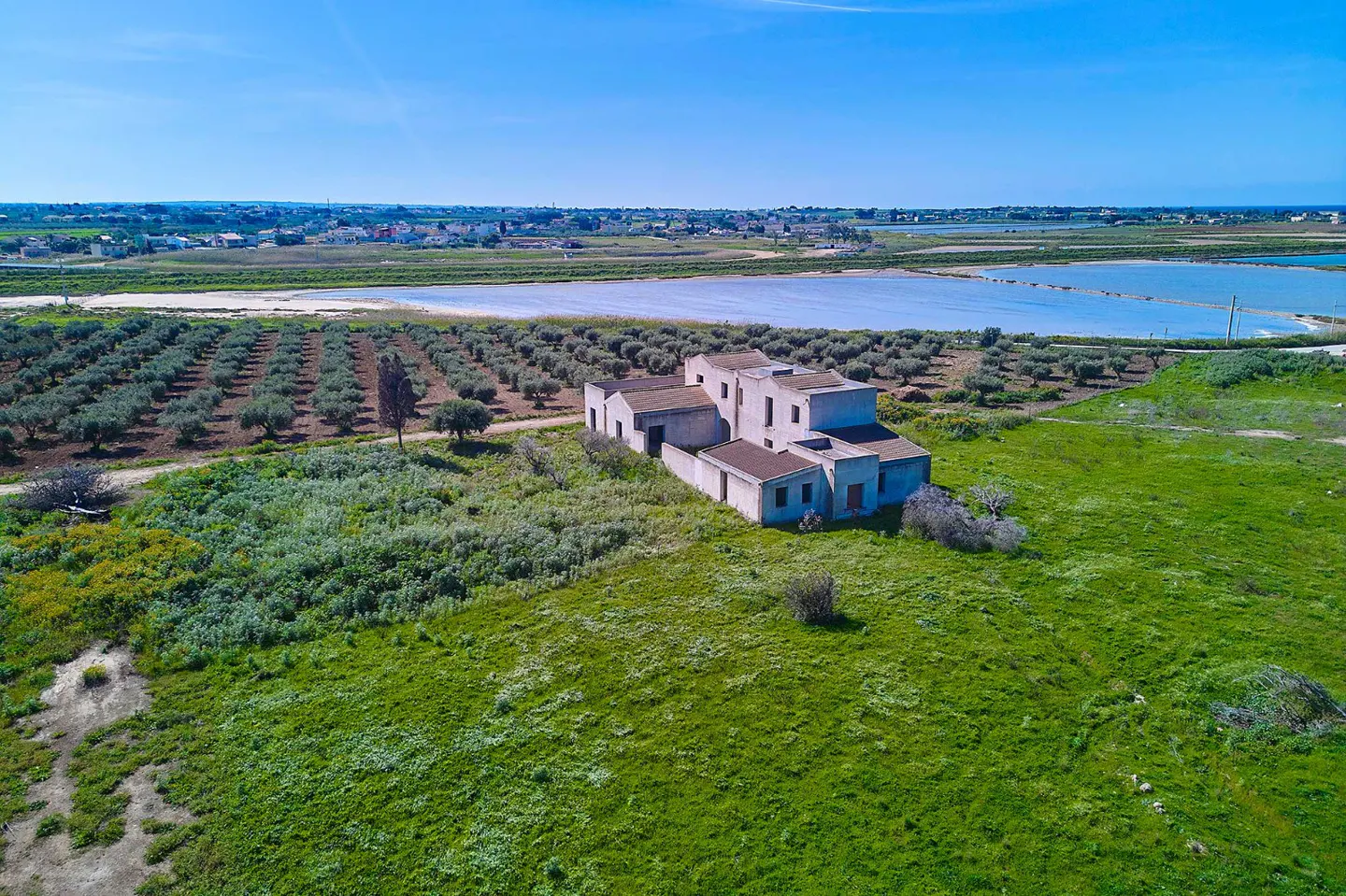 Aerial view of a white, multi-sectioned house surrounded by green grass, olive trees, and a blue sky. Salt flats are visible in the background.