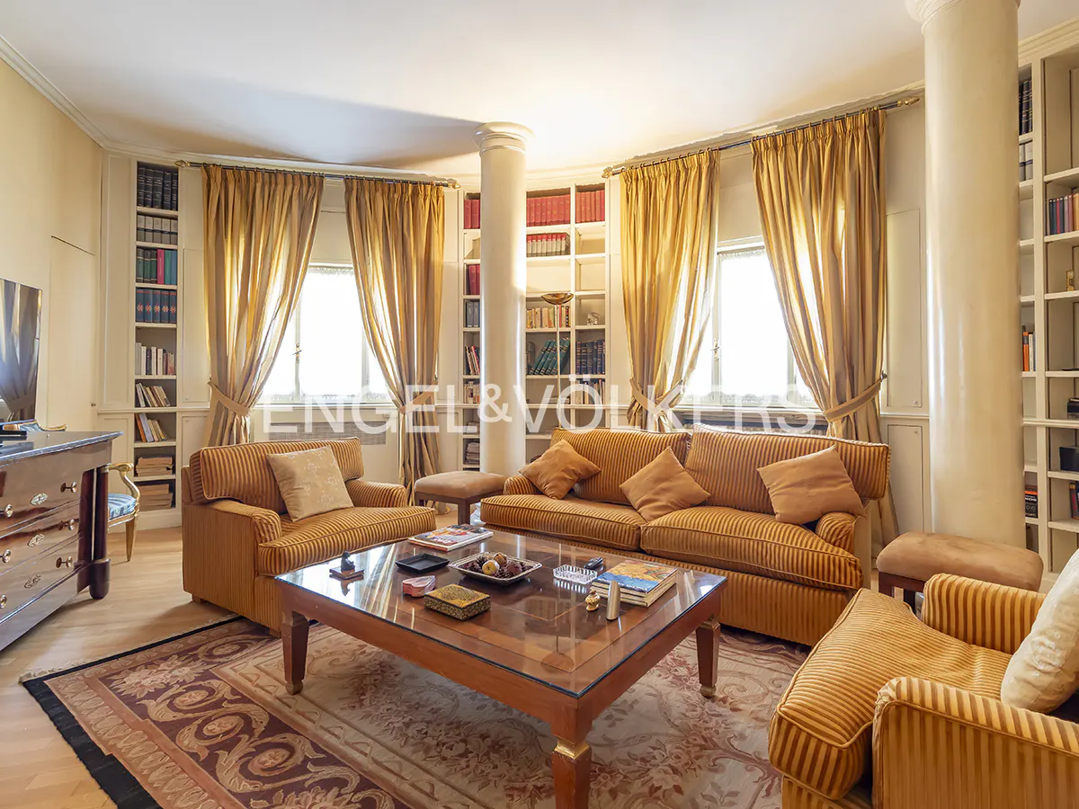 Living room with striped sofas, a glass-topped coffee table, and floor-to-ceiling bookshelves. Gold curtains frame the windows.