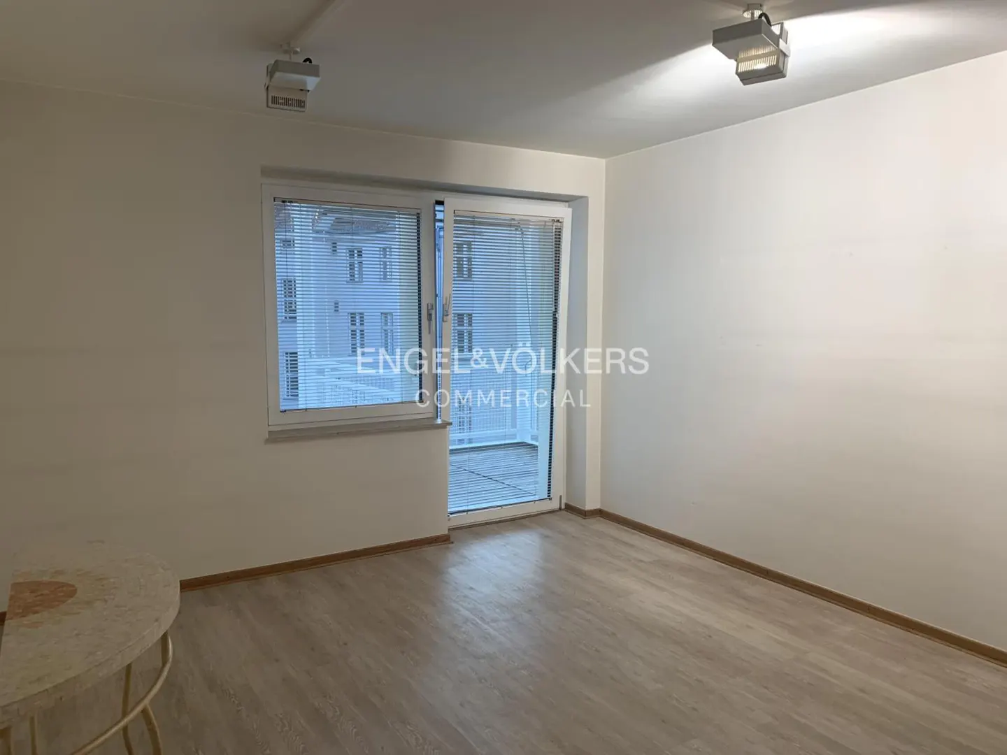 Empty room with light wood floors, white walls, and a glass door to a balcony. Engel & Völkers logo on the door.