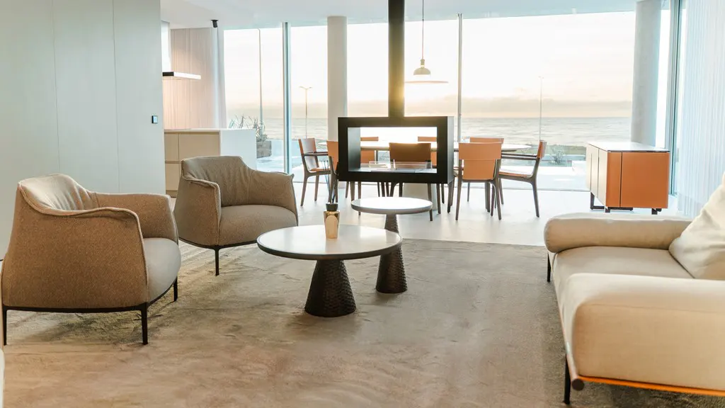 Bright, modern living room with beige armchairs, sofa, and round tables. A black fireplace hangs in front of a large window with an ocean view.