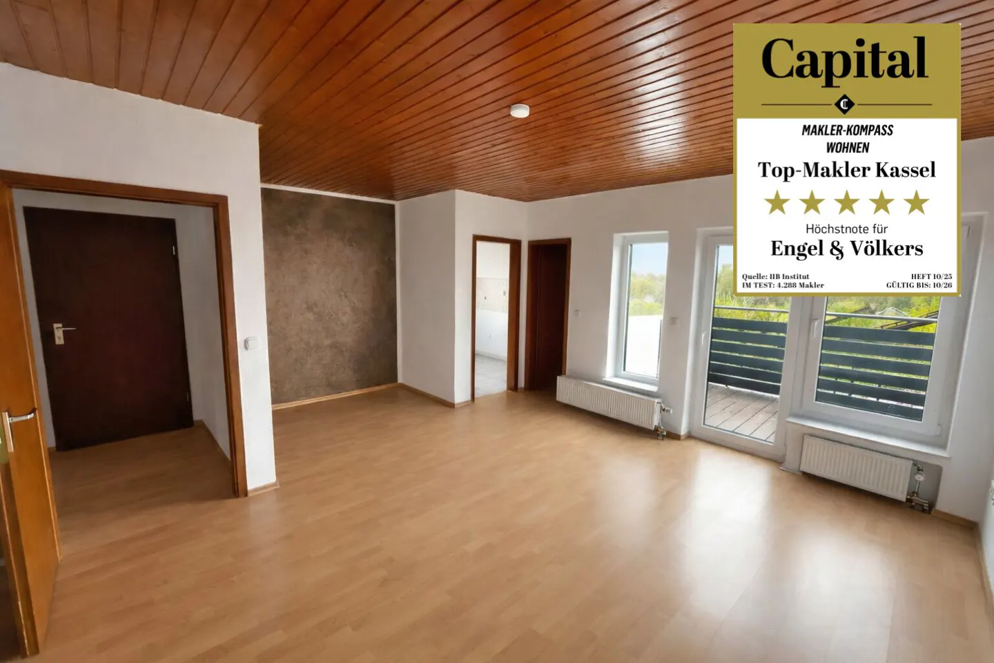 Bright, empty room with wood floors, a brown wood ceiling, and white walls. A balcony is visible through the windows. A "Top-Makler Kassel" award is displayed.