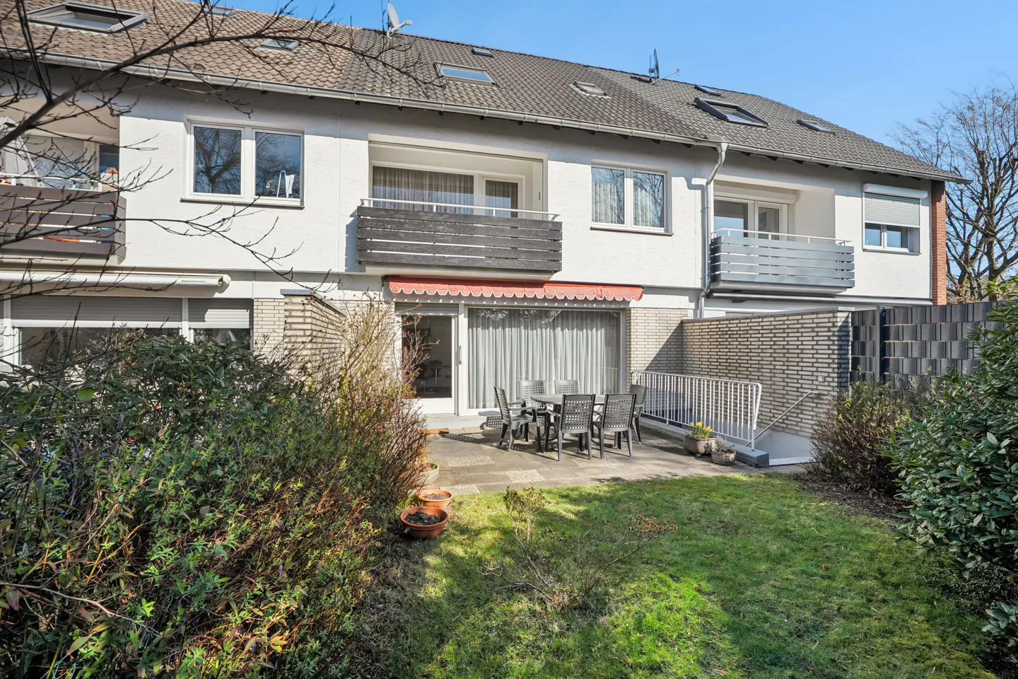 Backyard view of a three-story white townhouse with balconies and a patio set on a sunny day.