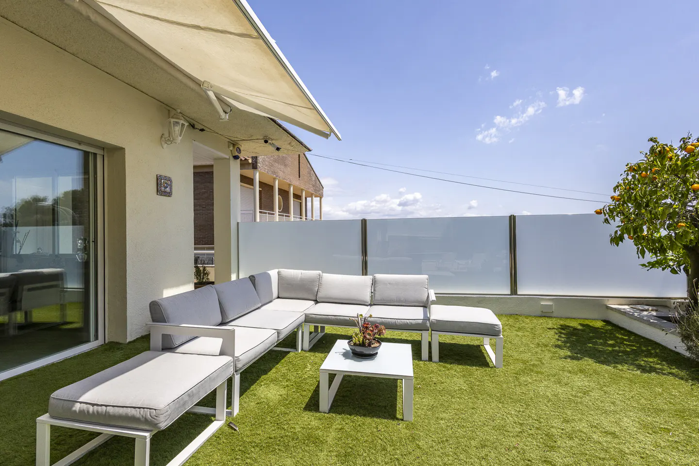 Sunny rooftop terrace with artificial grass, white sectional sofa, coffee table, and a small orange tree under a canopy.
