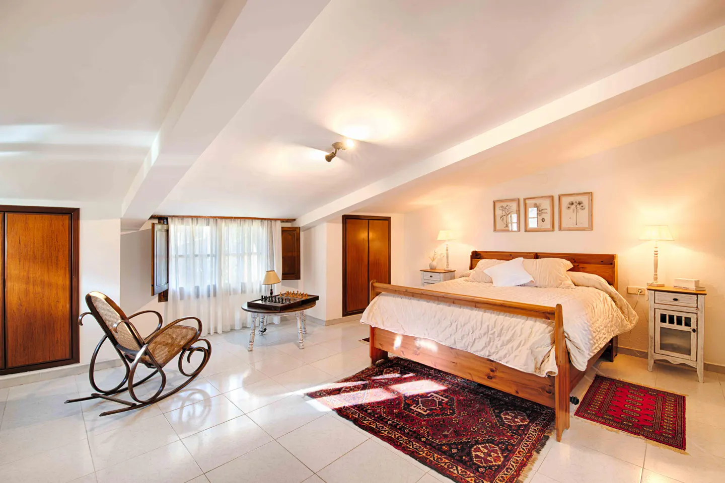 Bedroom with a wooden bed, white linens, bedside tables with lamps, and a rocking chair near a window.
