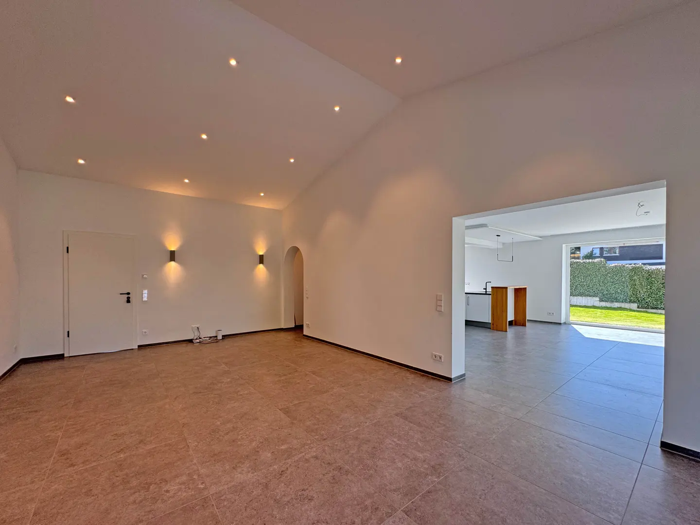 Bright, empty room with brown tile floor, white walls, and recessed lighting. An open doorway leads to a kitchen and a view of a green lawn.