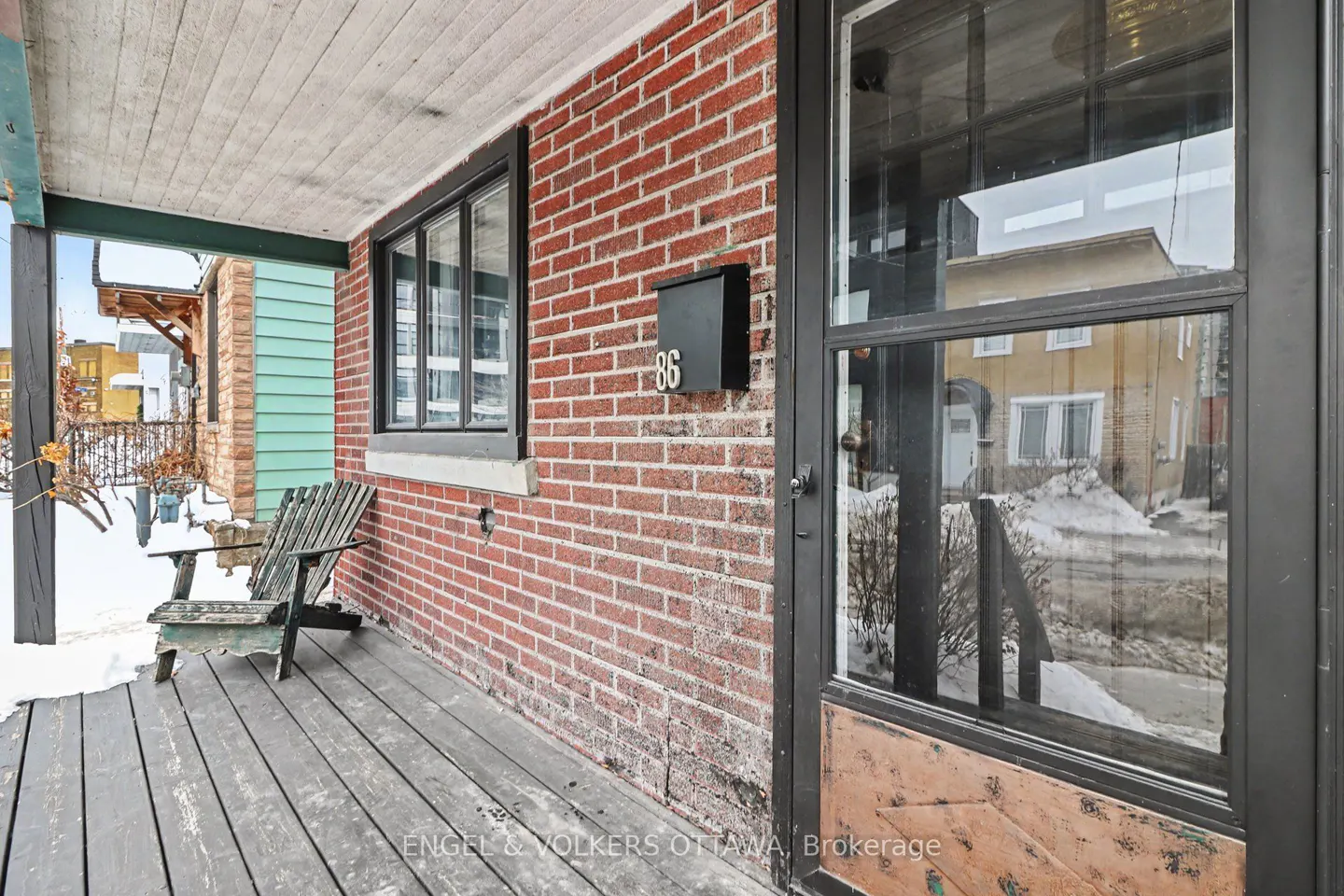 Covered porch of a red brick house with a black door, window, and mailbox labeled "86". A wooden chair sits on the porch. Snow is visible.