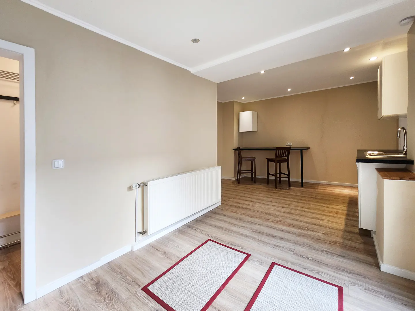 A bright, open apartment space with light wood floors, beige walls, and a small kitchen area. A white radiator is on the wall, and two small rugs are on the floor.
