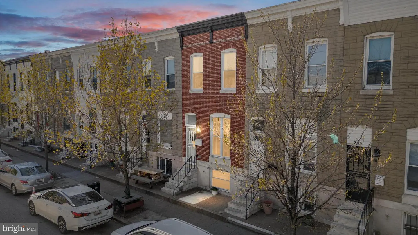 Row of colorful brick row houses with trees lining the street at dusk. Cars are parked along the curb.
