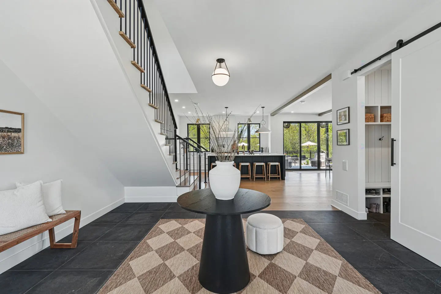 A bright foyer with black tile floors, a staircase, and a black table with a white vase. A sliding barn door leads to a mudroom.
