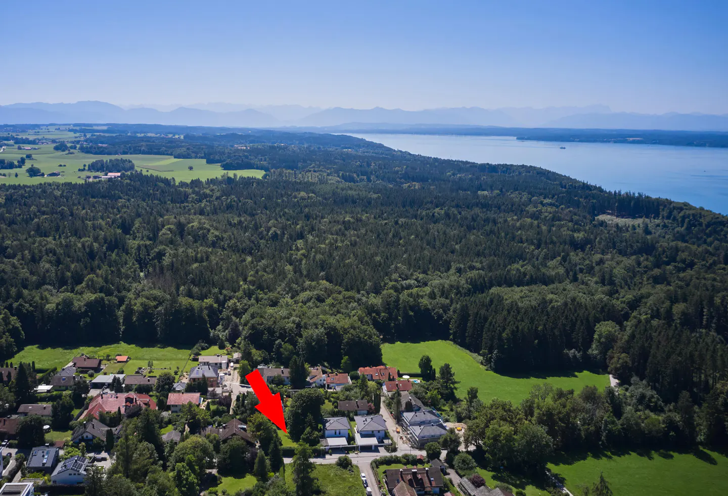 Aerial view of a property with a red arrow pointing to it, surrounded by trees, houses, and a lake in the background.