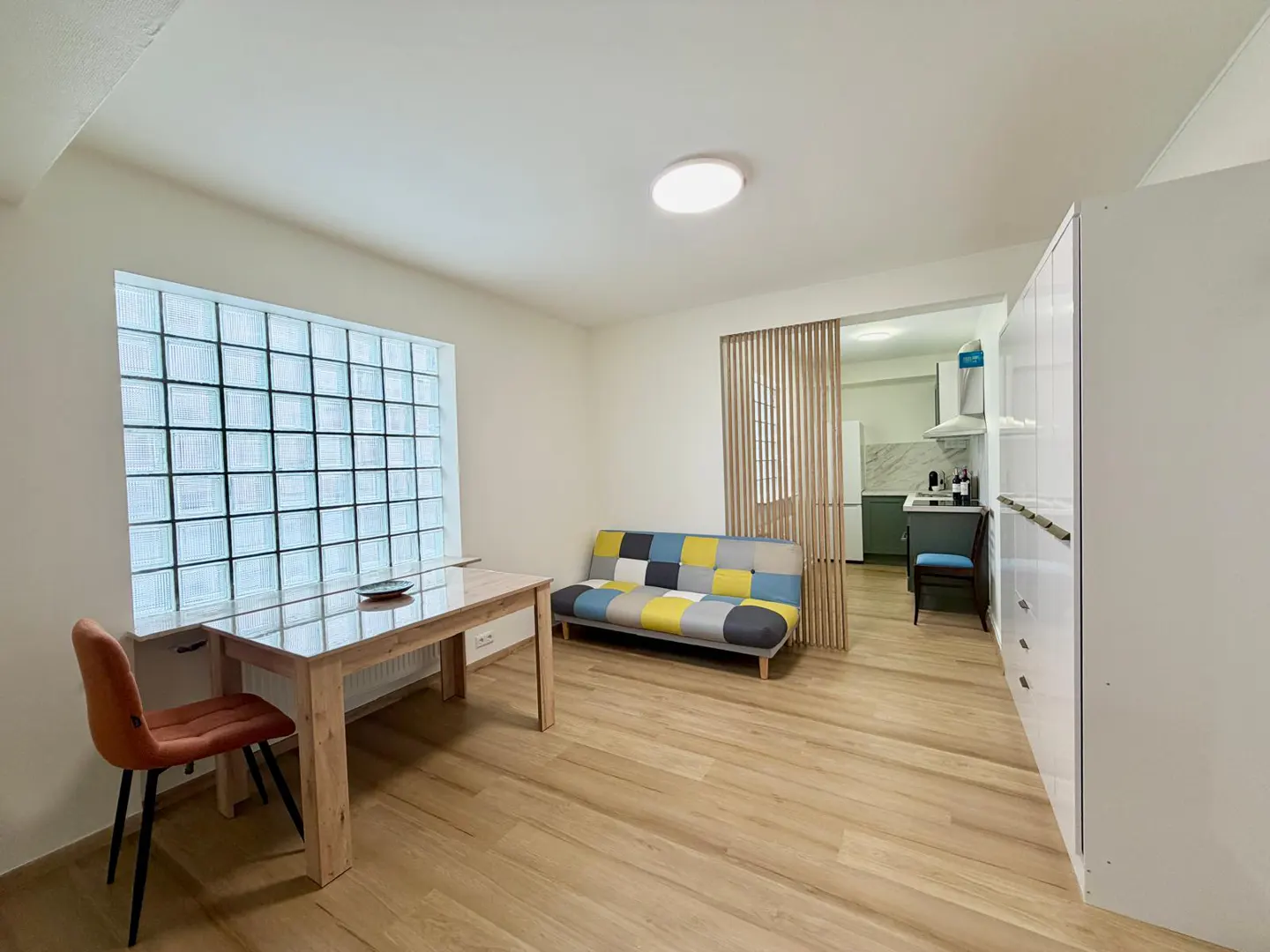 Modern studio apartment with a glass block window, wooden dining table, colorful sofa, and a view into a small kitchen.