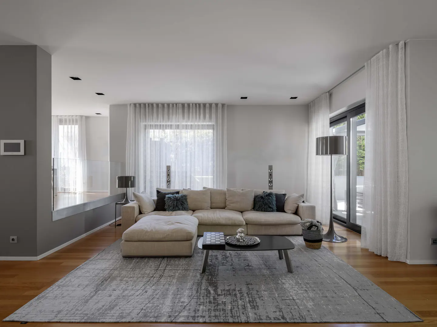 Modern living room with a beige sectional sofa, gray rug, and black coffee table. White curtains filter light from large windows.