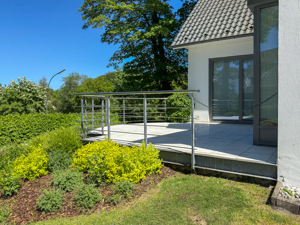 Modern white house with a tiled patio, stainless steel railing, and lush green garden under a clear blue sky.