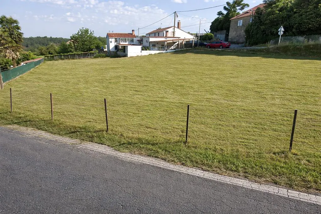 A grassy lot for sale, with a road in the foreground and houses in the background.