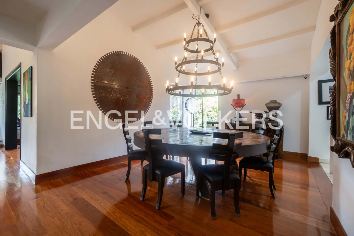 A dining room with a round wooden table, black chairs, and a large chandelier. A decorative metal disc hangs on the wall.