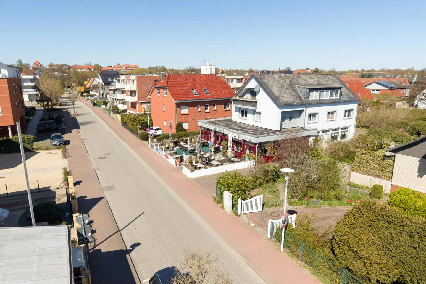 Aerial view of a quiet street with houses, trees, and a restaurant with outdoor seating on a sunny day.