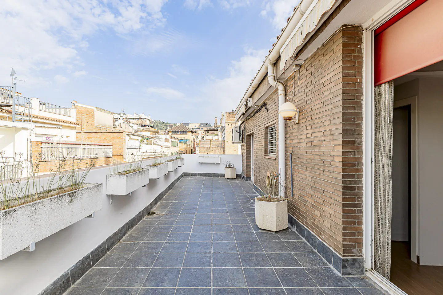 A rooftop terrace with gray tiles, white planters, and a brick building. Cityscape visible in the background under a blue sky.