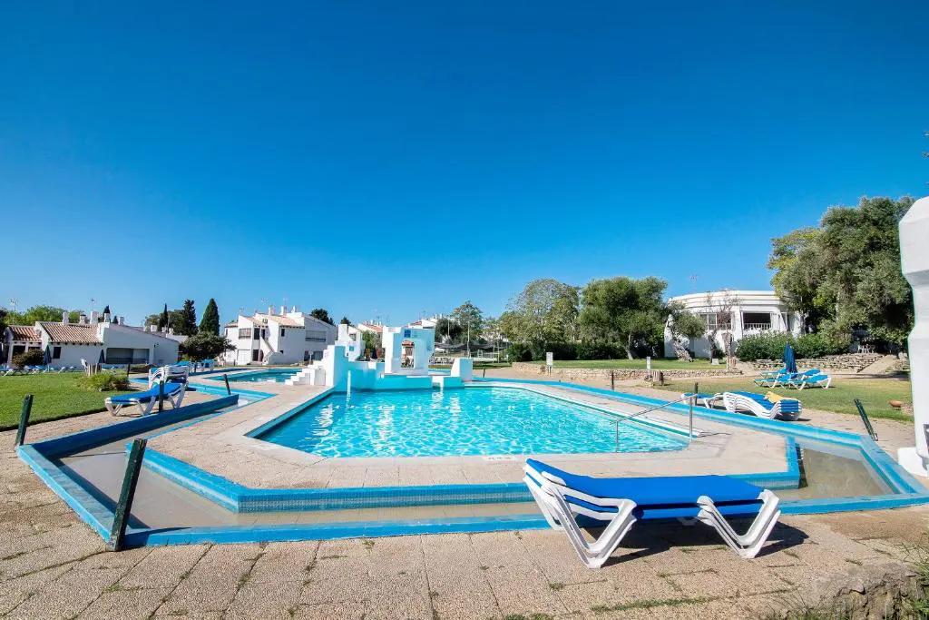 Outdoor pool with blue water and white lounge chairs. White buildings and green grass surround the pool under a clear blue sky.