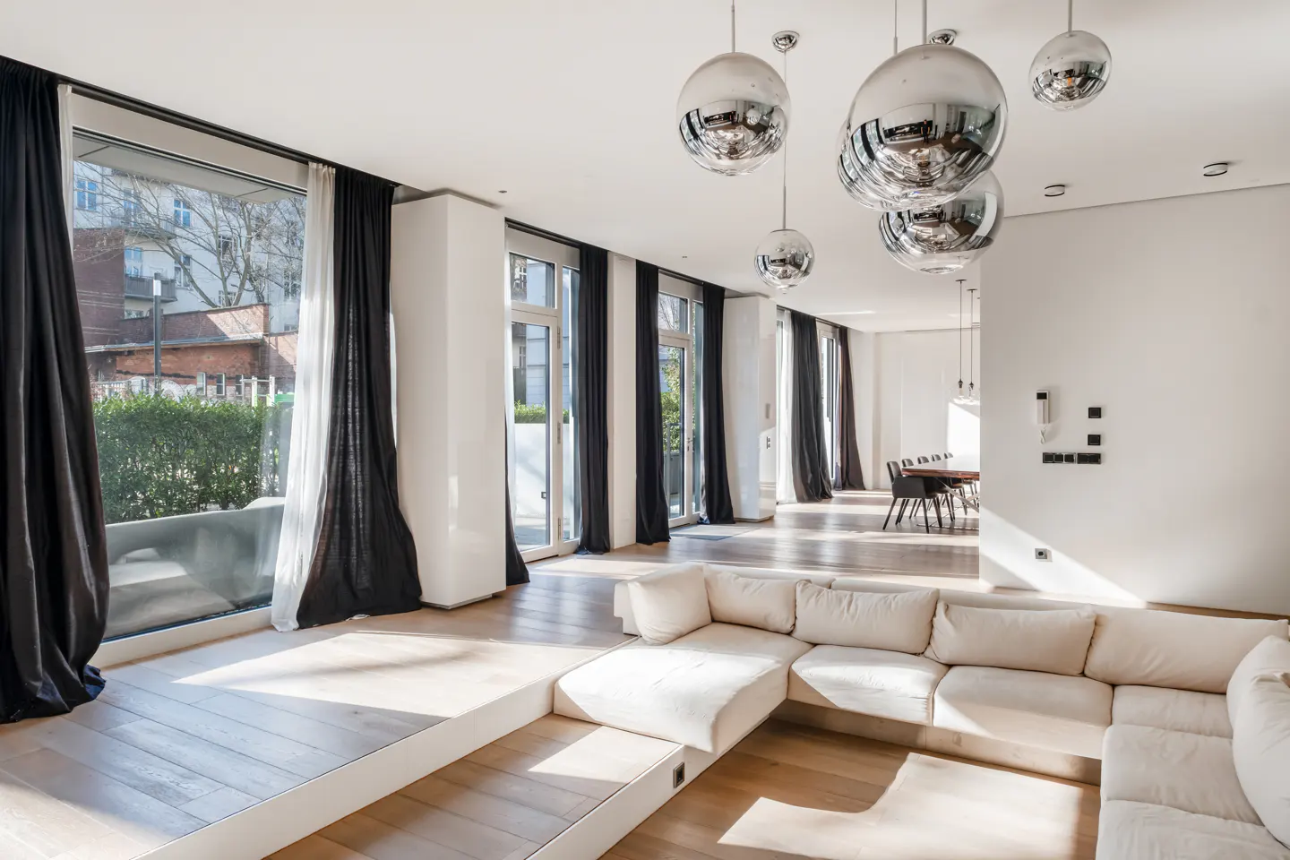 Bright, modern living room with a white sectional sofa, wood floors, and black curtains. Large windows offer a view of greenery. Silver, spherical pendant lights hang from the ceiling.