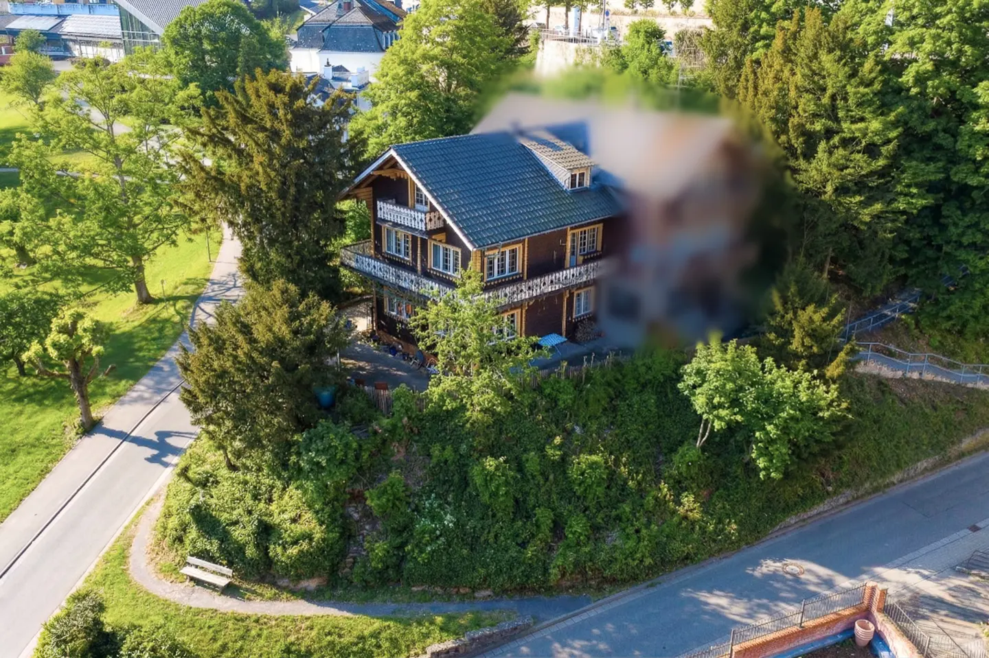 A three-story brown house with a blue roof and white trim, surrounded by green trees and foliage.