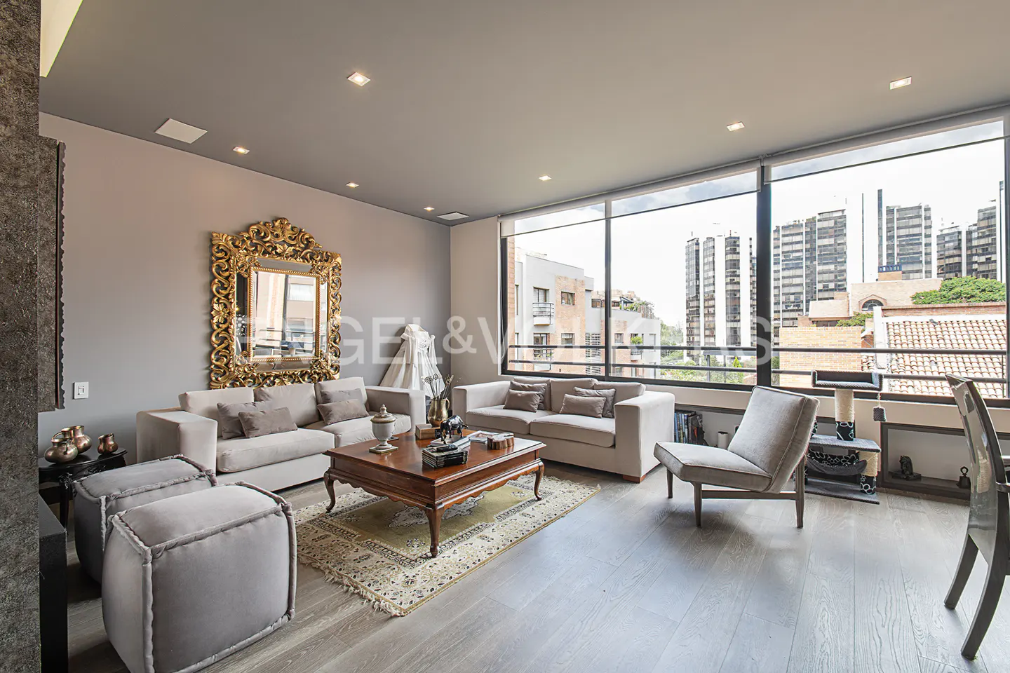 A modern living room with gray sofas, a wooden coffee table, and a large gold-framed mirror. A large window shows a city view.