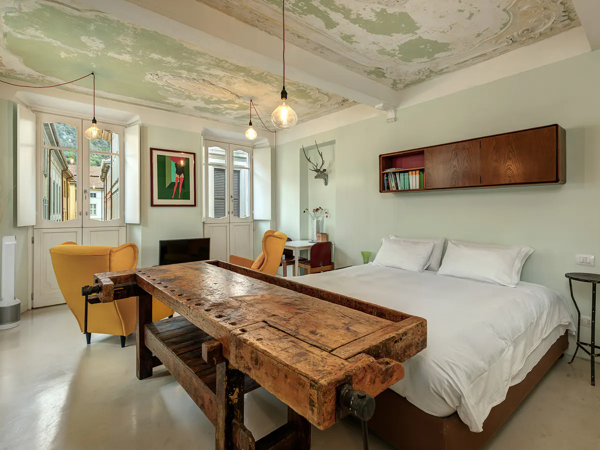 Bedroom with a rustic workbench in front of a white bed. Yellow chairs and a view through white doors add color. The ceiling has a distressed, decorative finish.