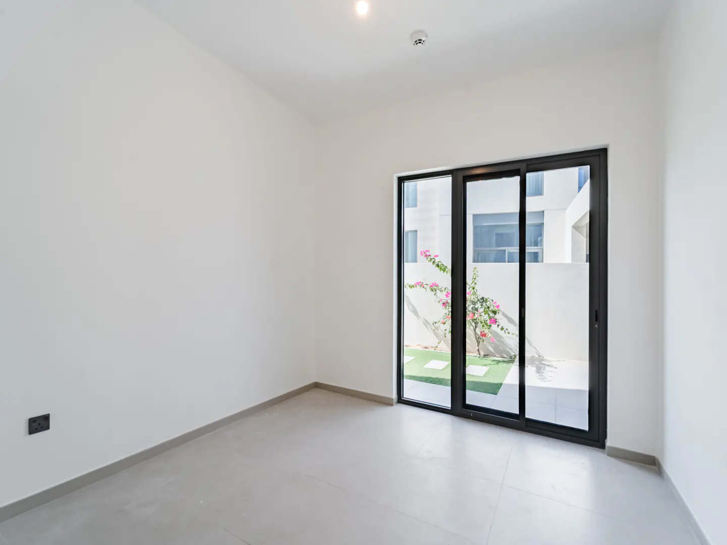 Empty room with white walls, light tile floors, and black sliding glass doors opening to a patio with pink flowers.