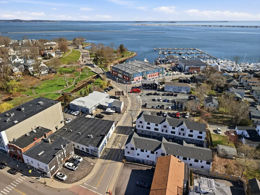 Aerial view of a coastal town with a marina, buildings, and a park under a blue sky.