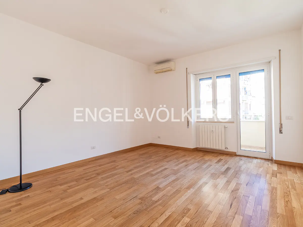 Bright, empty room with wood floors, white walls, and a black floor lamp. Balcony doors let in natural light. Engel & Völkers logo on wall.