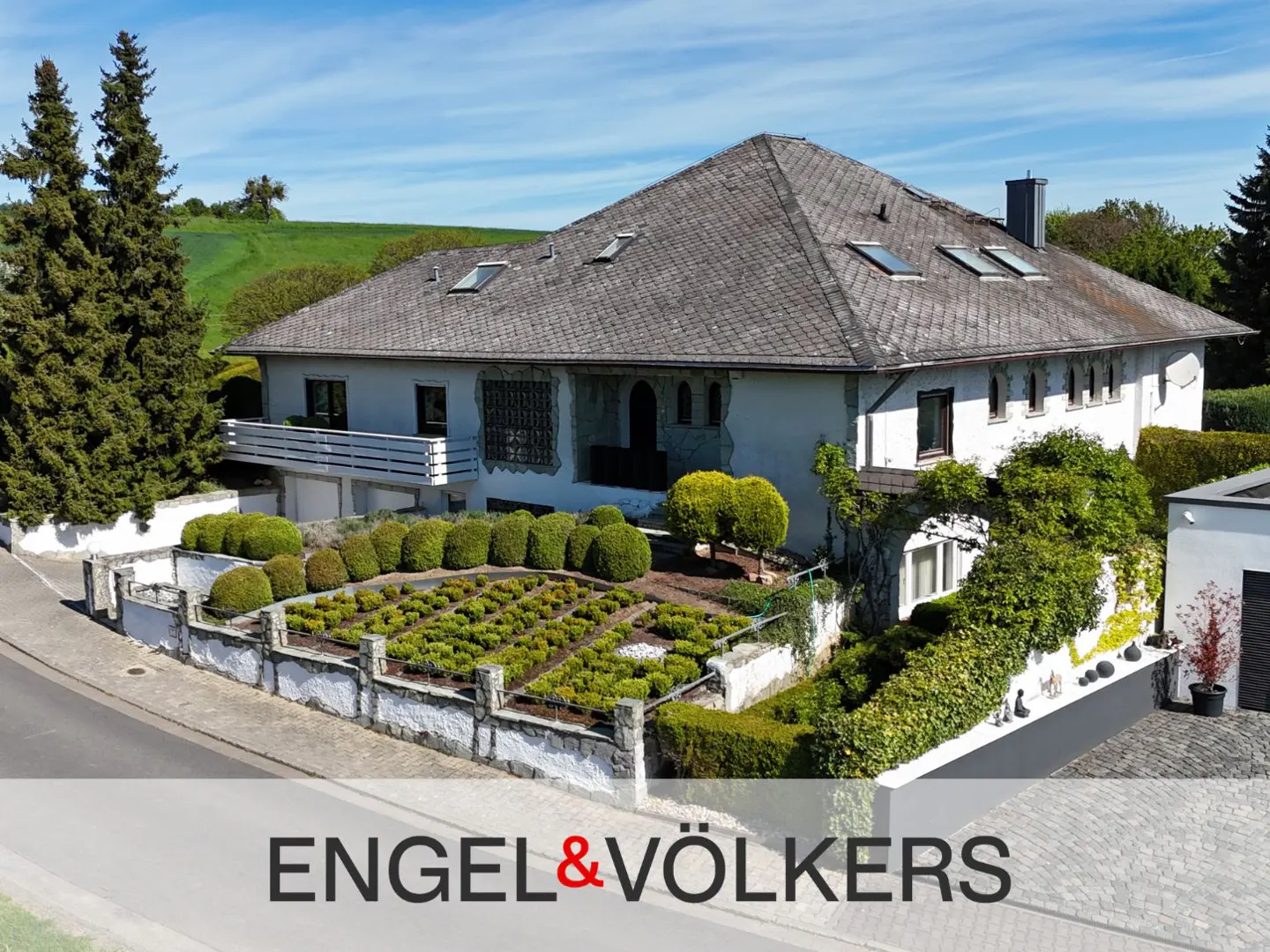 Exterior of a two-story white house with a gray roof and manicured green garden. Engel & Völkers logo at the bottom.