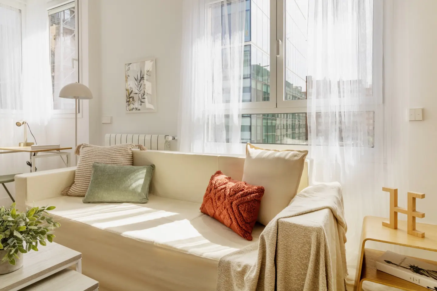Bright living room with a beige sofa, decorative pillows, and sheer white curtains. A wooden side table holds a decorative "H" and a book.