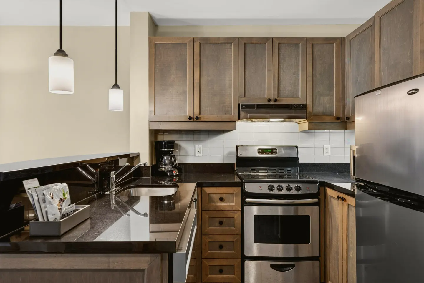 A kitchen with stainless steel appliances, dark countertops, and wood cabinets. Two pendant lights hang over the counter.