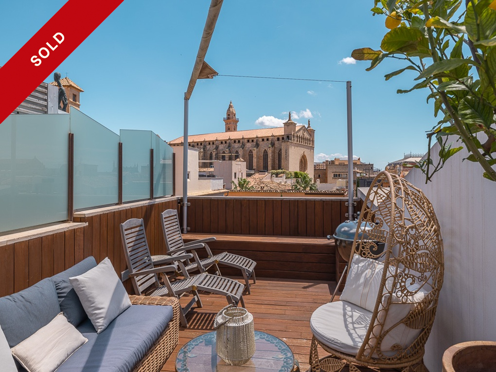 Rooftop terrace with wooden deck, lounge furniture, and a wicker egg chair, overlooking a historic stone church.