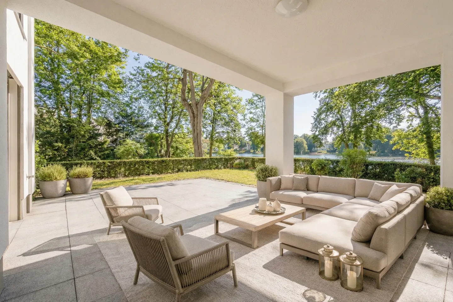 Covered patio with beige outdoor furniture, including a sectional sofa, chairs, and a coffee table. Green trees and a lake are visible in the background.