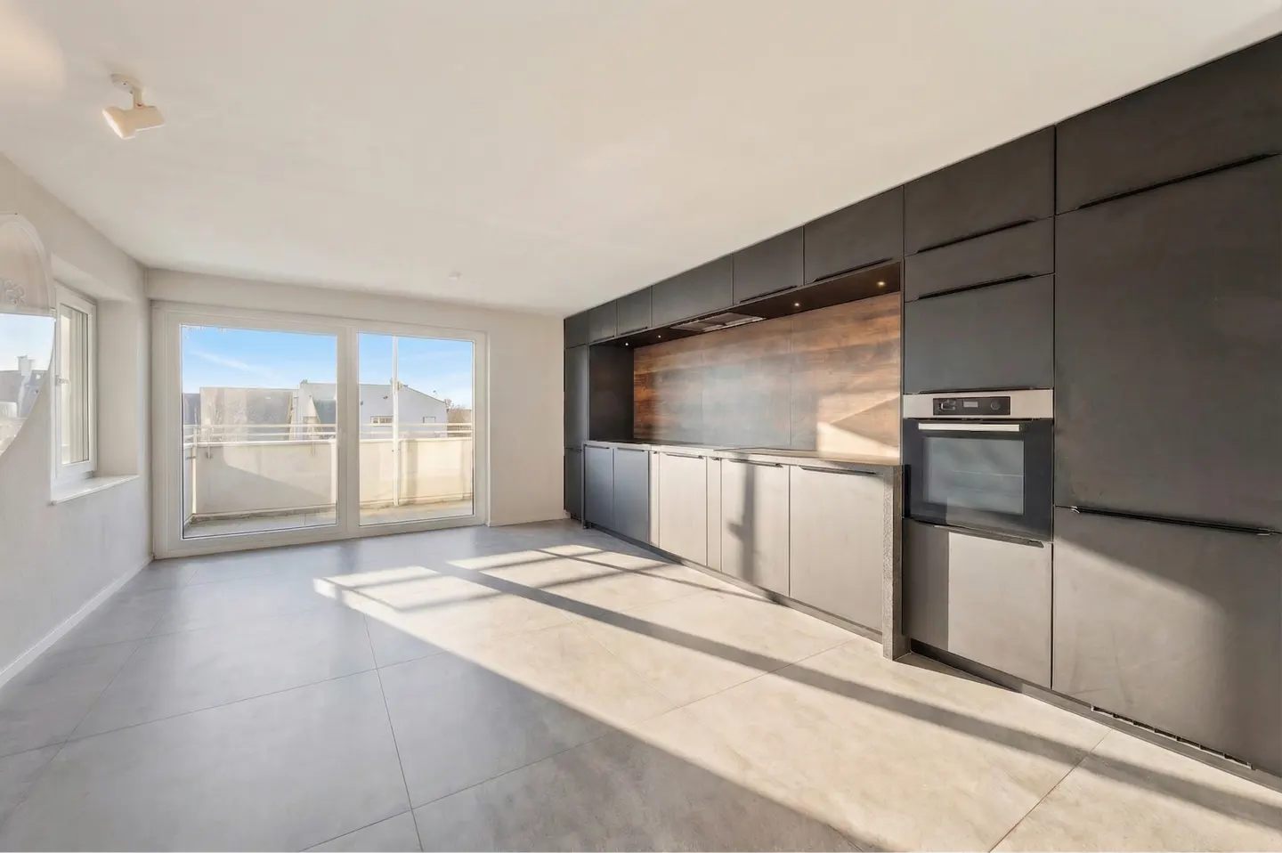 A modern kitchen with gray cabinets, stainless steel oven, and a sliding glass door to a balcony. Sunlight streams across the tiled floor.
