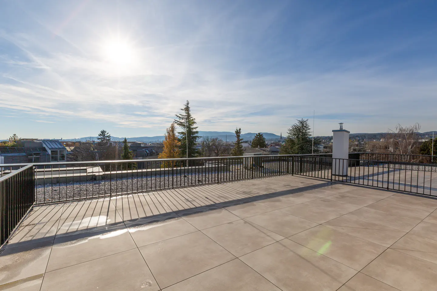 Wide rooftop patio with gray tiles and black railings, overlooking a cityscape under a blue sky with scattered clouds and a bright sun.