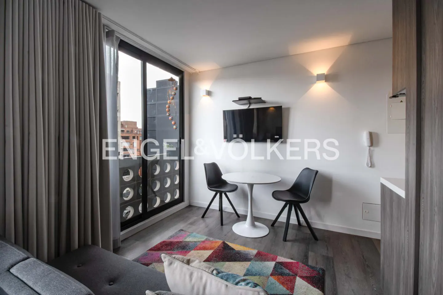 Living room with gray sofa, colorful rug, and a white table with two black chairs. A TV is mounted on the wall.