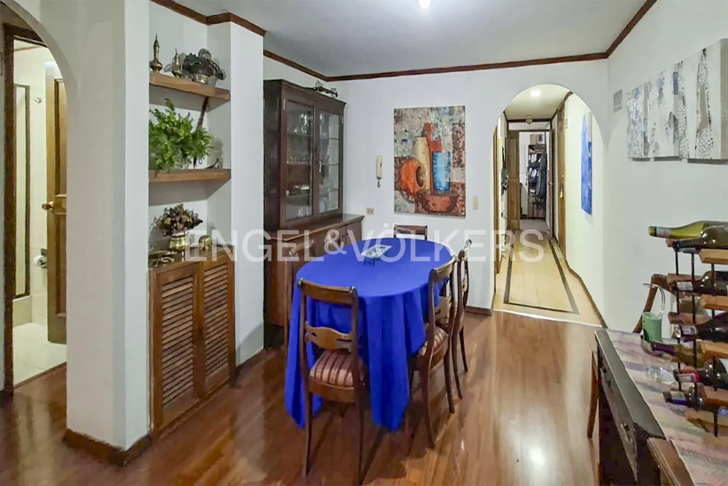 Dining room with a blue-clothed oval table, wooden chairs, a hutch, wall art, and a wine rack on hardwood floors.
