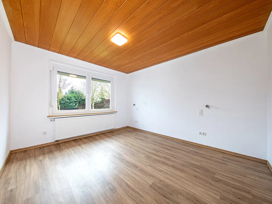 Bright, empty room with wood-look floors, white walls, and a wood plank ceiling. A window overlooks green trees.
